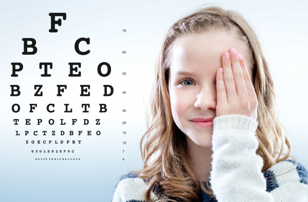 A young girl covering her left eye with her hand while undergoing an acuity test and a Snellen chart in the background.