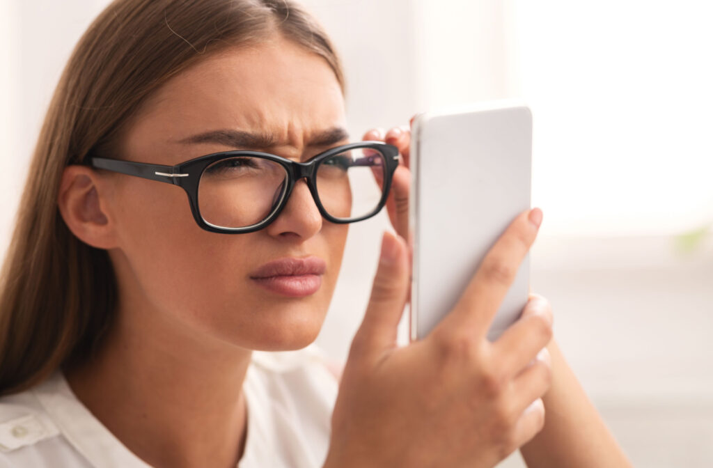 A woman with glasses holding her phone in one hand and squinting to read as she struggles with myopia.