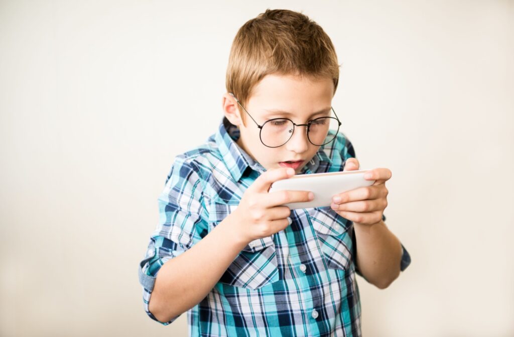 A child with glasses standing, holding a smartphone very close to his face