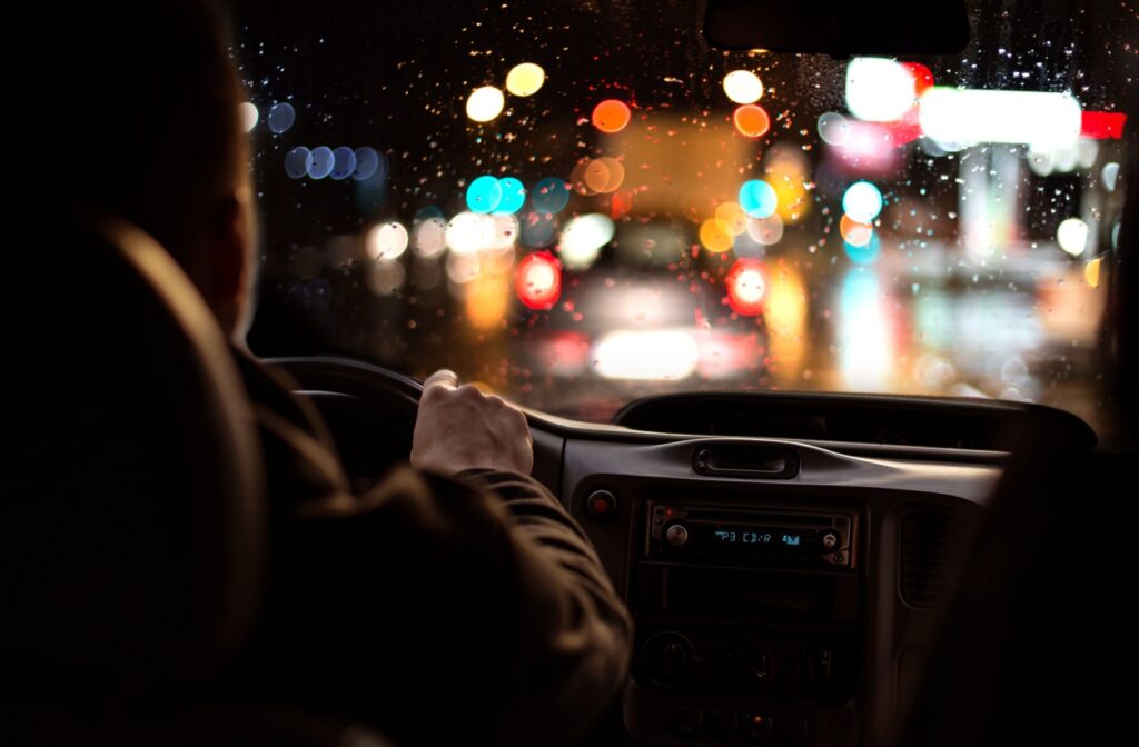 A silhouette of a person driving a car at night, seeing halos and glare from surrounding traffic.