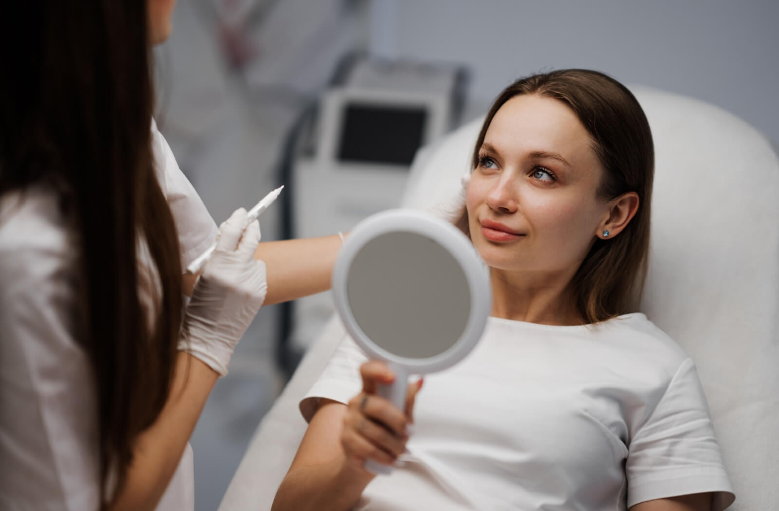 A person sitting in a treatment chair, holding a mirror and smiling at a practitioner who is discussing IPL treatment.